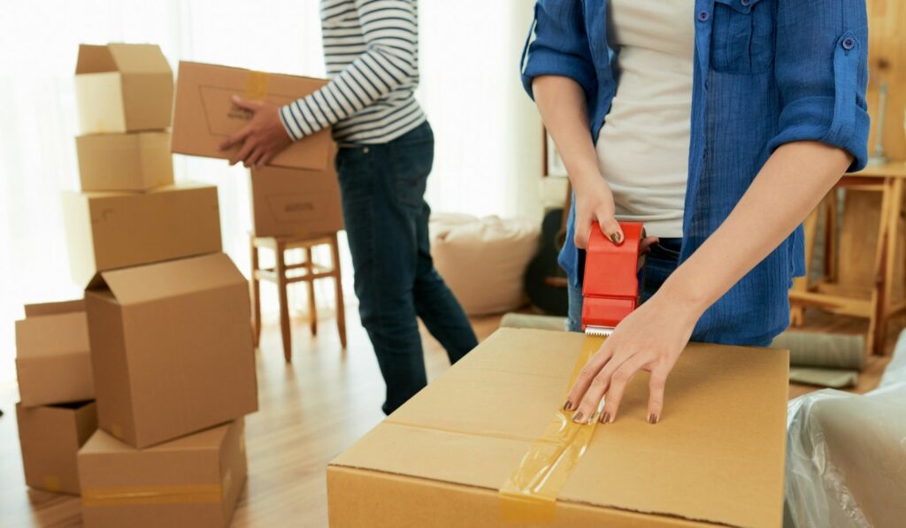 Woman taping moving boxes with a man moving boxes in the background.