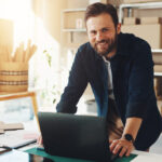 A smiling man standing at a desk in front of his small business inventory.