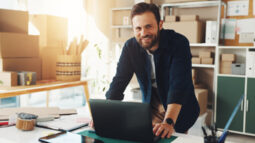 A smiling man standing at a desk in front of his small business inventory.