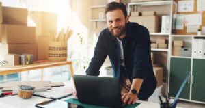 A smiling man standing at a desk in front of his small business inventory.