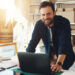 A smiling man standing at a desk in front of his small business inventory.