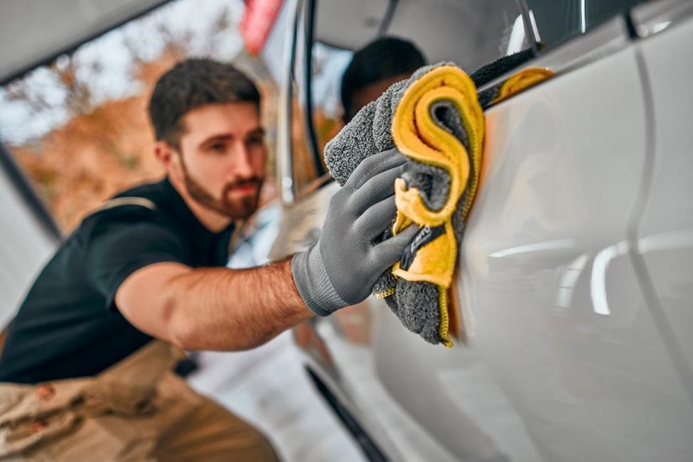 A man wearing gloves uses a gray and yellow microfiber cloth to polish the rear passenger side door of a white car.