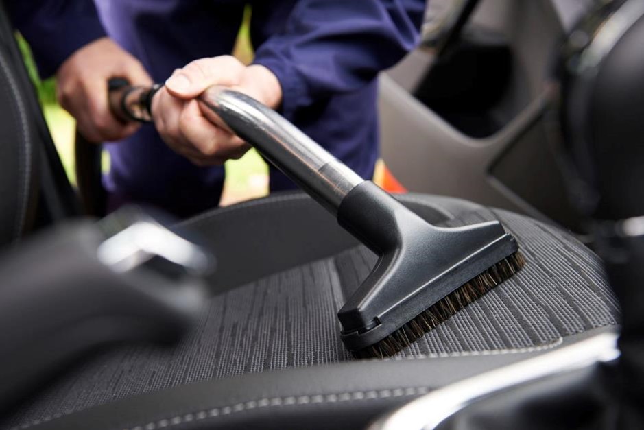 A man uses a vacuum cleaner with a brush attachment to clean the black fabric seat of a vehicle. 