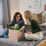 Two dorm roommates sitting on a bed next to a packed box.