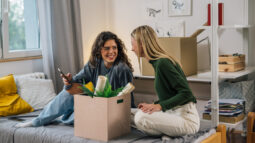 Two dorm roommates sitting on a bed next to a packed box.