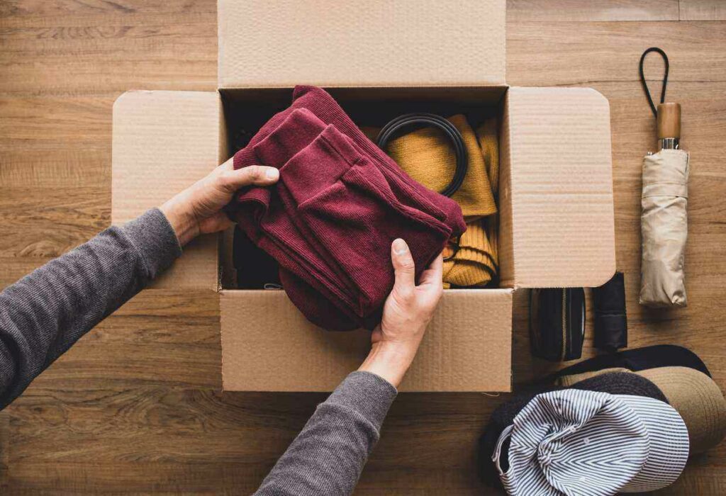 A man packs a red sweatshirt into a small cardboard box. An umbrella and ball caps sit nearby