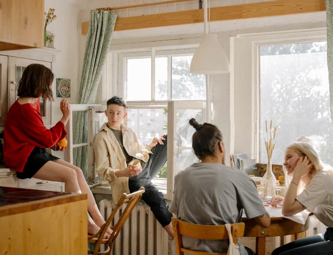 A group of four friends sit in a kitchen, on counters, and in the window as they talk and catch up.