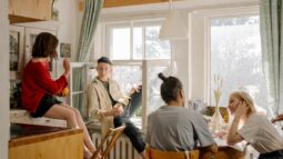 A group of four friends sit in a kitchen, on counters, and in the window as they talk and catch up.