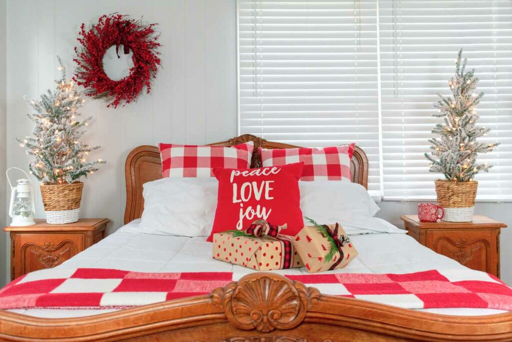 A wooden sleigh bed is decorated with red checkered bedding, presents, a pillow that reads “peace, love, joy” and two silver trees on the side tables