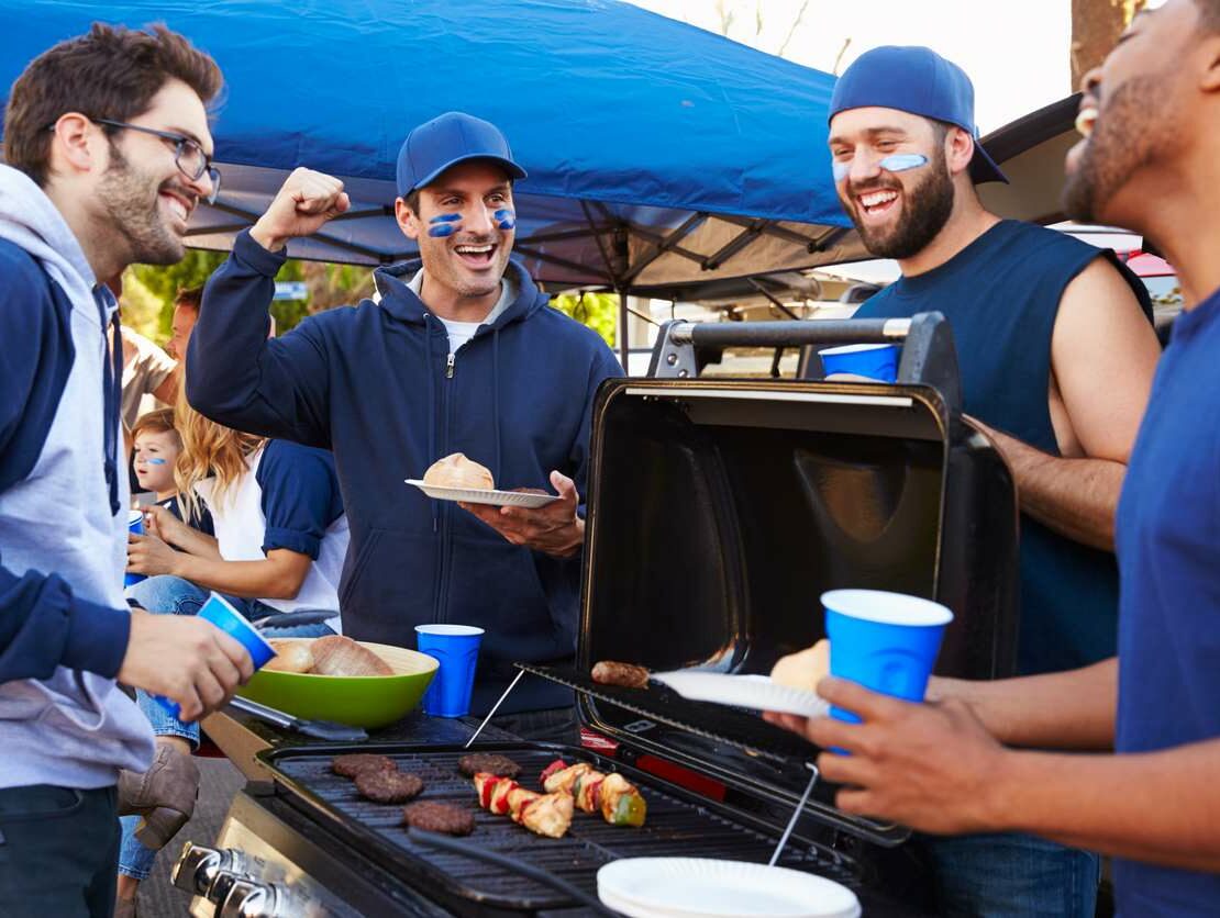 A group of friends stands around a grill at a sports tailgate while cooking food and talking