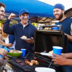 A group of friends stands around a grill at a sports tailgate while cooking food and talking