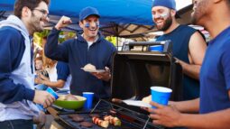 A group of friends stands around a grill at a sports tailgate while cooking food and talking
