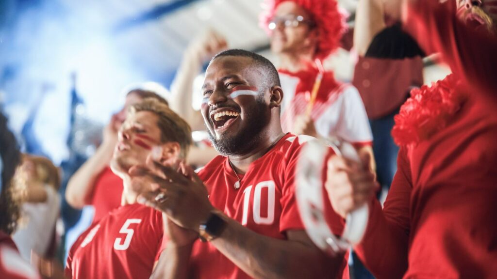 A man wearing a red jersey with red and white face paint cheers at a sports game