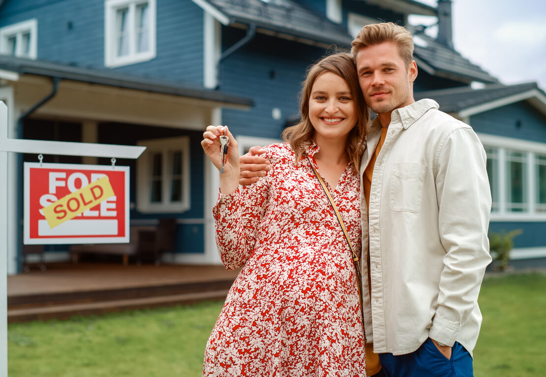 A couple standing in front of a house and a "sold" sign.