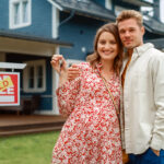 A couple standing in front of a house and a "sold" sign.