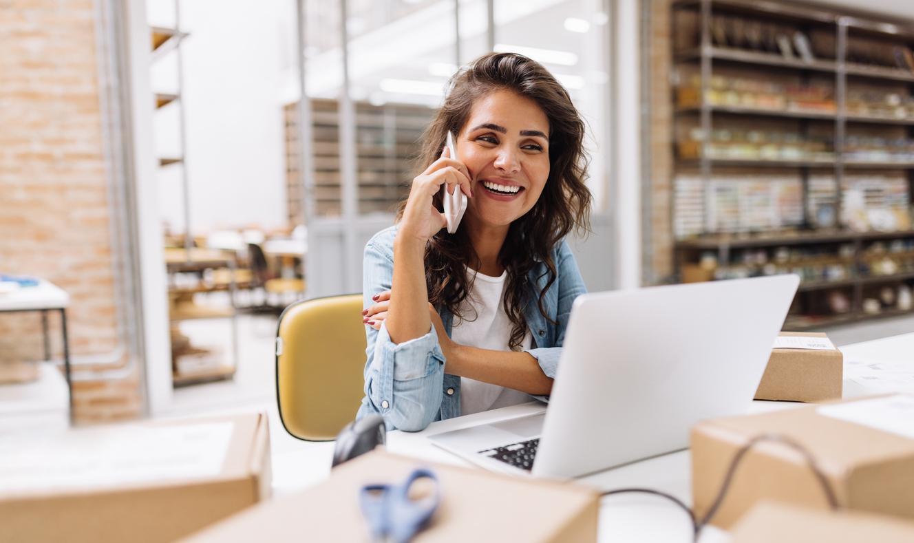 A smiling woman surrounded by boxes and talking on the phone.