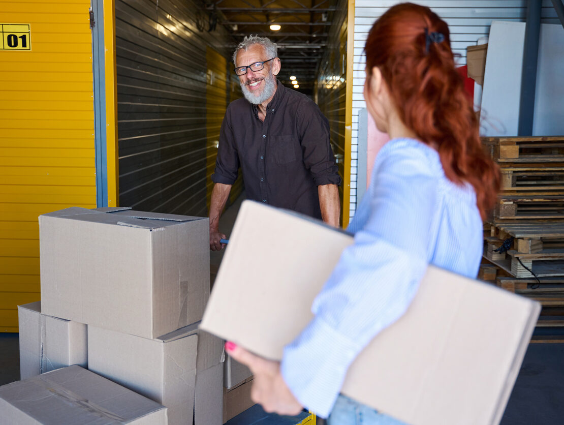 A man smiling at a woman as he pushes a cart packed with boxes at a self storage facility.