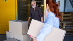 A man smiling at a woman as he pushes a cart packed with boxes at a self storage facility.