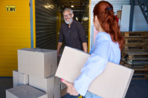 A man smiling at a woman as he pushes a cart packed with boxes at a self storage facility.