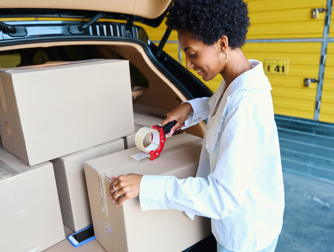 A smiling woman taping a box shut on the edge of her open car trunk.