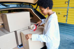 A smiling woman taping a box shut on the edge of her open car trunk.