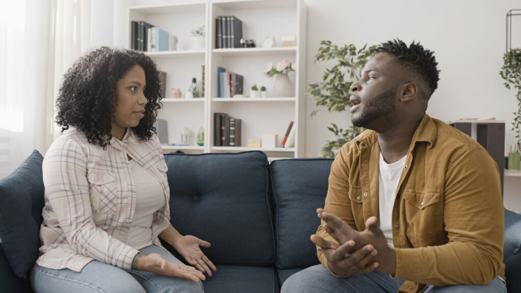 A man and woman sitting on the couch communicating their feelings to one another.