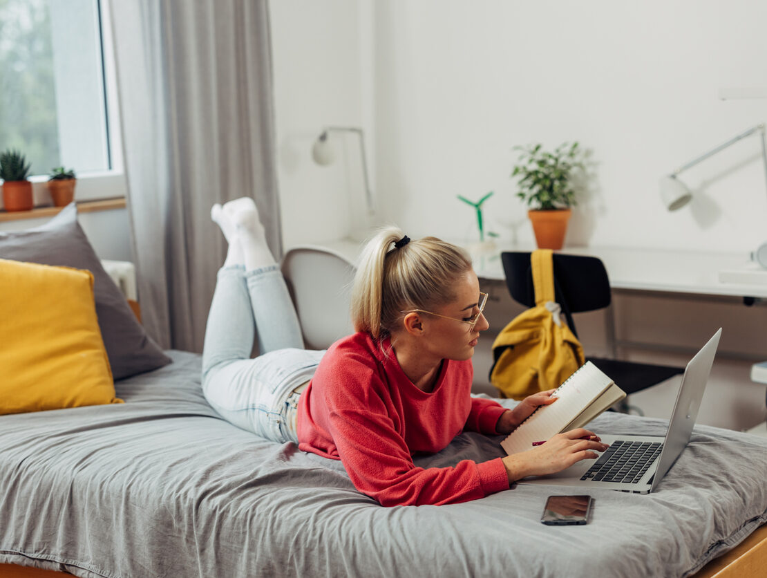 A college student sitting on the bed and taking notes from her laptop in her freshman dorm room.