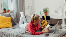 A college student sitting on the bed and taking notes from her laptop in her freshman dorm room.