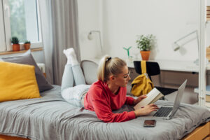 A college student sitting on the bed and taking notes from her laptop in her freshman dorm room.