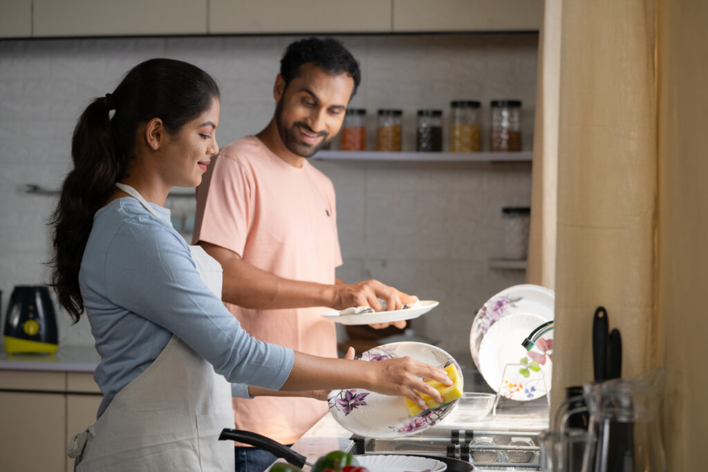 A smiling man and woman washing dishes together in their kitchen sink.