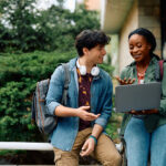 Two young college students smiling and laughing while looking at a laptop.