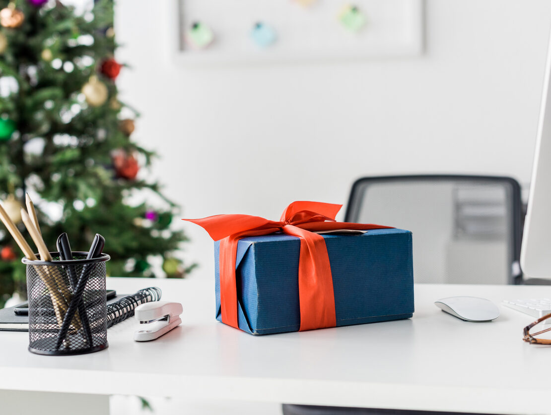 A blue present wrapped in a red bow sitting on a white office desk, with a Christmas tree in the background.