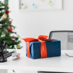 A blue present wrapped in a red bow sitting on a white office desk, with a Christmas tree in the background.