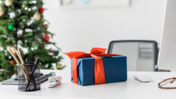 A blue present wrapped in a red bow sitting on a white office desk, with a Christmas tree in the background.