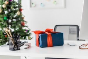 A blue present wrapped in a red bow sitting on a white office desk, with a Christmas tree in the background.