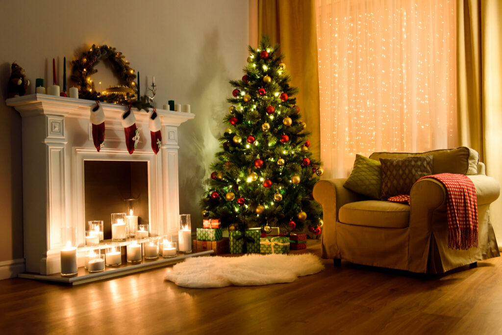 A classic Christmas tree by a fireplace decorated with candles, a wreath, and other mantle decorations.