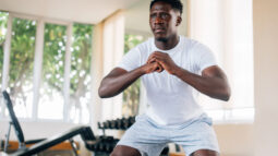 A man squats in his home gym with a dumbbell rack and adjustable bench in the background.