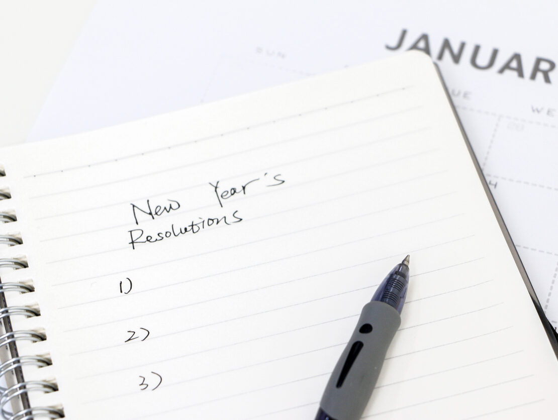 A pen rests on a notebook where someone has written “New Year’s Resolutions” and a blank list numbered one through three.