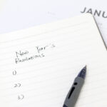 A pen rests on a notebook where someone has written “New Year’s Resolutions” and a blank list numbered one through three.