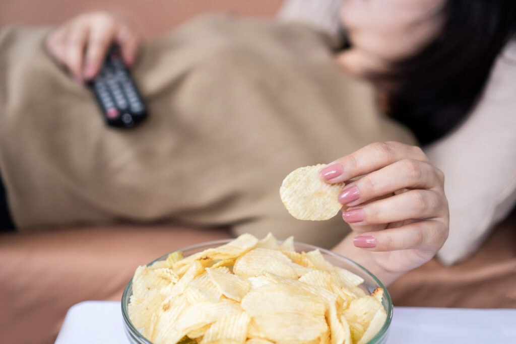 An adult woman lying down watching TV and eating chips instead of working out for the new year.