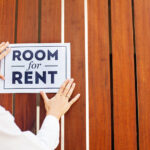 A woman placing a sign on a fence that says “Room for Rent.”