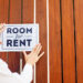 A woman placing a sign on a fence that says “Room for Rent.”