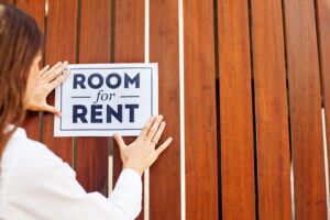 A woman placing a sign on a fence that says “Room for Rent.”