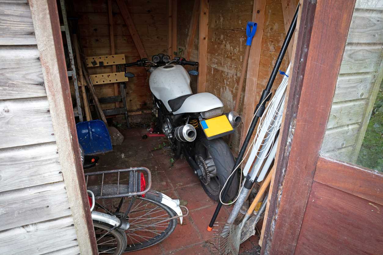 A motorcycle is stored inside a small wooden shed with garden tools and a bicycle nearby.