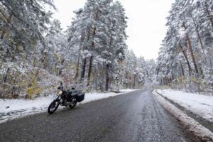 A black motorcycle parked on a snowy forest road surrounded by tall pine trees covered in fresh winter snow.