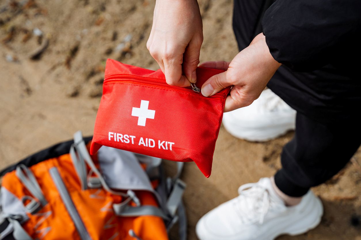 A person holding a red first aid kit with a white cross on it, next to a backpack on sandy ground.