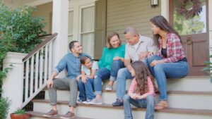 A family sitting together on a front porch, smiling and talking on the steps of a home surrounded by plants and decor.