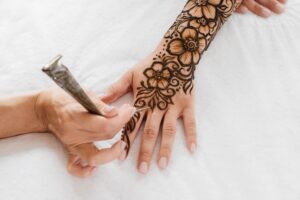 A close-up of a henna artist applying an intricate floral mehndi design to a person's hand and forearm