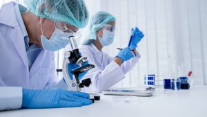 Two laboratory researchers wearing protective equipment conduct testing with a microscope and sample tubes in a bright, clean lab.